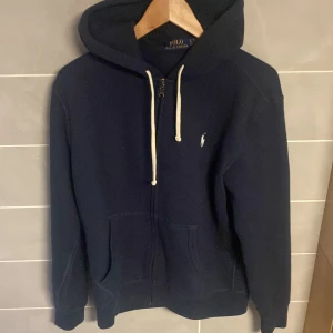 Mörkblå hoodie från Polo Ralph Lauren - Säljer en klassisk mörkblå hoodie från Polo Ralph Lauren med vit broderad logga på bröstet. Tröjan har dragkedja, huva med vita snören och två fickor framtill. Perfekt för en avslappnad stil.  Nypris runt 2000kr. 100% ÄKTA