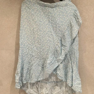 Ljusblå blommig omlottkjol - Säljer en ljusblå omlottkjol med små vita blommor och volangdetalj längs kanten. Kjolen har en luftig känsla och är perfekt för varmare dagar. Passar dig som gillar romantisk och söt stil.