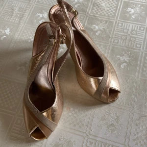 Guldiga klackskor med öppen tå - Säljer ett par eleganta guldiga klackskor med öppen tå och smal rem runt hälen. Skorna har en glansig yta med diskret mönster och en beige detalj framtill. Perfekta för dig som vill sticka ut med stil.