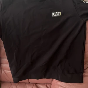 Svart sweatshirt från EA7 Emporio Armani - Svart sweatshirt från EA7 Emporio Armani med tryckt logga på bröstet och längs ena ärmen. Tröjan har rund halsringning och långa ärmar. Perfekt för en sportig och stilren look.