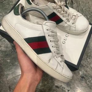 Gucci vita sneakers med gröna och röda ränder - Säljer ett par klassiska vita sneakers från Gucci med gröna och röda ränder på sidorna och grön detalj på hälen. Skorna har snörning och rund tå. Perfekta för dig som gillar stilrena och ikoniska märken. 