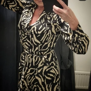 Zebramönstrad omlottklänning med bälte - Säljer en lång zebramönstrad omlottklänning i svart och beige med lång ärm och knytbälte i midjan. Klänningen har v-ringad halsringning och markerad midja för en snygg siluett. Perfekt för dig som vill sticka ut med ett djurmönster! Pris: 600kr