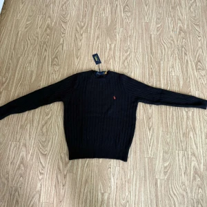 Svart stickad tröja från Polo Ralph Lauren - Klassisk svart stickad tröja från Polo Ralph Lauren med diskret röd logga broderad på bröstet. Tröjan har rund halsringning och långa ärmar, samt ribbade muddar vid ärmslut och nederkant. Perfekt för en stilren look.