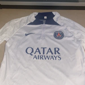 Vit PSG långärmad fotbollströja från Nike - Säljer en vit Paris Saint-Germain fotbollströja från Nike med blå detaljer och klubbmärke på bröstet. Tröjan har korta ärmar och Qatar Airways som sponsortryck på framsidan. 