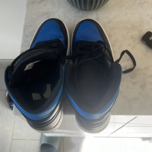 Nike Air Jordans i svart och blå - Säljer ett par Nike Air Jordans high-top sneakers i svart och blå med vit sula. Skorna har klassisk snörning och den ikoniska swoosh-loggan på sidan. Perfekta för dig som gillar streetwear och vill sticka ut med en färgstark look.