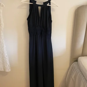 Mörkblå långklänning med spetsdetalj från VILA - Säljer en mörkblå långklänning från VILA i storlek 34. Klänningen har v-ringning, är ärmlös och har en vacker spetsdetalj i midjan. Perfekt för dig som vill ha en elegant och stilren look. Använd en gång.