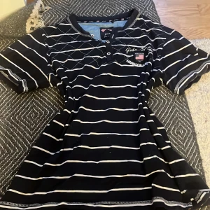 Svart och vitrandig t-shirt från J.F.K - Svart t-shirt med vita ränder från J.F.K. Modellen har korta ärmar, knappar vid halsen och tryckt logga samt amerikansk flagga på bröstet. Snygg och sportig stil med klassiska detaljer.