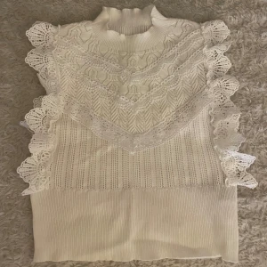 Vit stickad topp med spetsvolanger - Säljer en vit stickad topp med hög krage och dekorativa spetsvolanger längs axlarna och framtill. Toppen har ett ribbat material och fina virkade spetsdetaljer. Den är lite mindre i storleken men den är i toppen skick och är helt ny