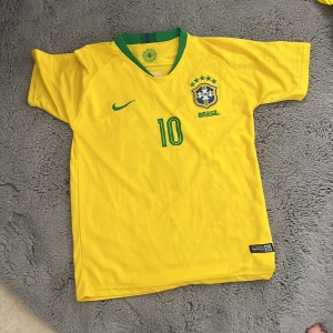 Brasilien fotbollströja Neymar JR 10 - Gul Brasilien fotbollströja med gröna detaljer, nummer 10 och Neymar JR på ryggen. Nike-logga på framsidan och Brasilien-emblem med stjärnor. Klassisk design med rund hals och korta ärmar.