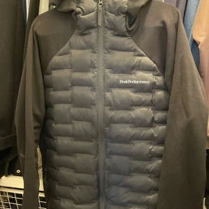 Svart hybridjacka från Peak Performance - Säljer en svart hybridjacka från Peak finns en mindre reva med vadderad framsida och rygg samt mjuka ärmar och sidor. Jackan har huva och dragkedja framtill. Perfekt för lager-på-lager och aktiva dagar