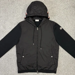 moncler cardigan | as najs | ev byte - Snygg svart jacka från Moncler med huva och dragkedja. Jackan har stickade ärmar och quiltad kropp i syntetmaterial. Huvan har en randig detalj i rött, vitt och blått. Kan även bytes