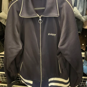 Mörkblå trackjacka från GANT - Säljer en mörkblå trackjacka från GANT med vit dragkedja och vita ränder vid midjan. Jackan har hög krage och GANT-logga broderad på bröstet. Perfekt för en sportig och avslappnad stil.