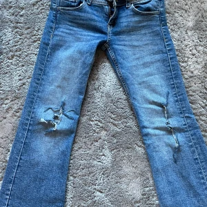 Blå slitna jeans från Gina Tricot - Ett par blå jeans från Gina Tricot med utsvängda  ben och slitna detaljer på båda benen. Jeansen har klassisk femficksdesign och normal passform. Perfekta för en avslappnad och trendig look.