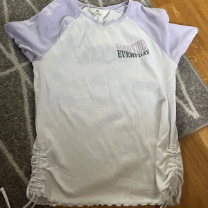 Vit och ljuslila t-shirt från H&M - Vit t-shirt från H&M med ljuslila ärmar och texten 'GOOD VIBES EVERYDAY' på bröstet. Kortärmad modell med rynkade detaljer och knyt i sidorna. 