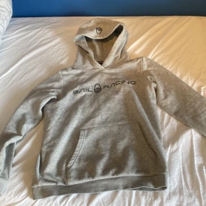 Grå hoodie från Sail Racing - Säljer en grå hoodie från Sail Racing med svart logga och text på bröstet samt logga på huvan. Tröjan har magficka och ribbade muddar. Perfekt för en avslappnad stil.