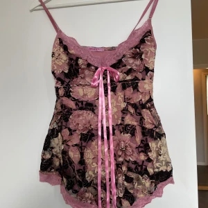 Blommig spetslinnetopp med rosett - Säljer en romantisk blommig linnetopp i rosa och svart med spetsdetaljer och en söt rosa rosett framtill. Toppen har tunna axelband och är tillverkad i ett lätt, transparent material med blommönster och glittriga inslag.Fint skick,använd vid ett tillfälle.