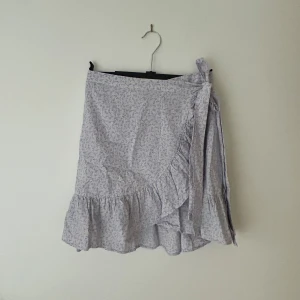 Ljuslila blommig omlottkjol från Vero Moda - Supersöt ljuslila omlottkjol från Vero Moda med små blommor i lila och gult. Kjolen har volangdetaljer längs kanten och knyts i sidan. Perfekt för varma dagar och ger en romantisk vibe.