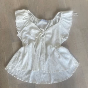 Vit blus med volang och knyt från 1/st - Söt vit blus från lager 157 med volangärmar och knyt framtill. Blusen har en lätt rynkad struktur och v-ringning. Storlek s