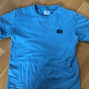 Blå t-shirt från C.P. Company - Säljer en blå t-shirt från C.P. Company med rund hals och ett svart broderat logomärke på bröstet. Klassisk passform och tillverkad i mjuk bomull. Perfekt för dig som gillar stilrena och snygga plagg.