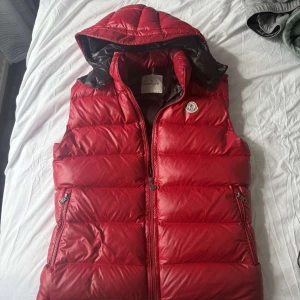 Röd dunväst från Moncler med huva - Säljer en röd dunväst från Moncler med avtagbar huva och dragkedja framtill. Västen har två fickor med dragkedja och klassisk Moncler-logga på bröstet. Insidan är svart och har serietidningsmönster. Perfekt för lager-på-lager-stil.