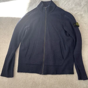 Mörkblå stickad cardigan tröja från Stone Island - Mörkblå stickad cardigan tröja från Stone Island med dragkedja framtill och ribbade muddar. Jackan har två framfickor och klassisk Stone Island-logga på ärmen. Perfekt för lager-på-lager och snygg till jeans.