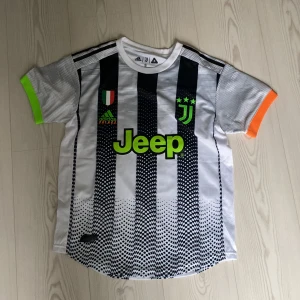 Juventus fotbollströja Ronaldo #7 - Juventus matchtröja med svarta och vita ränder, neongröna detaljer och tryck 'Ronaldo 7' på ryggen. Jeep-logga på bröstet, klubbmärke och italienska flaggan. Kortärmad modell med Climalite-material och färgade ärmslut.