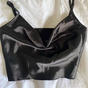 Svart satin croppad linnetopp från SHEIN - Säljer en svart croppad linnetopp i satin från SHEIN. Toppen har smala axelband och en snygg draperad urringning framtill. Perfekt att styla till olika outfits!