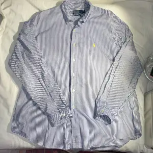 Säljer en stilren blå- och vitrandig skjorta från Polo Ralph Lauren som passar L. Skjortan har långa ärmar och en klassisk krage med knappar. Den är broderad med det ikoniska gula Polo-logotypen på bröstet. Perfekt för en elegant look.                                      Priset är inte hugget i sten.                                          Liknande plagg fins i på profilen!