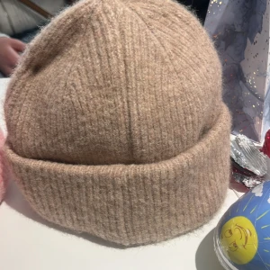 Beige stickad mössa med satin-foder - Snygg beige stickad mössa med mjukt satin-foder på insidan. Perfekt för att hålla värmen under kyliga dagar. Den ribbade designen ger en klassisk look.