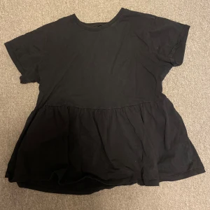 Svart blus med volangdetalj - Säljer en svart blus med korta ärmar och volangdetalj nedtill. Perfekt för en avslappnad stil. Blusen har en lös passform och är tillverkad i ett mjukt material.