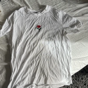 Vit t-shirt med ros från Shein - Säljer en vit t-shirt från Shein med en liten broderad ros på bröstet. T-shirten har en klassisk rund hals och korta ärmar. Perfekt för en enkel och stilren look.