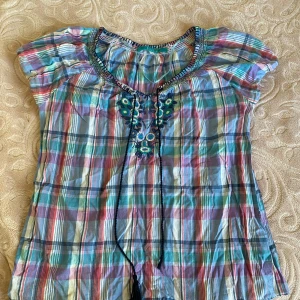 Rutig blus från RoseBud - Säljer en färgglad rutig blus från RoseBud i storlek M. Blusen har korta ärmar och ett vackert broderi med blommor framtill. Den har även en knytning vid halsen för extra detalj. Perfekt för sommaren!Den ser lite skrynklig ut i bilen men det är bara o stryka tröjan så blir den rak igen.💗