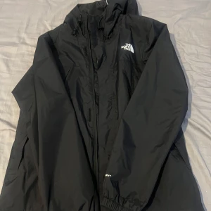The North Face regn/vindjacka  - Säljer en svart regn/vindjacka från The North Face med huva och dragkedja. Jackan har en klassisk design med logotyp på bröstet och ryggen. Perfekt för blåsiga dagar och lätt att packa med sig. Hör av er vid frågor. Kvitto finns!!!