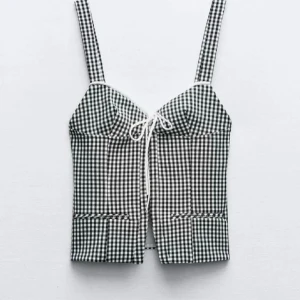 Svartvit rutig korsettopp - Snygg svartvit rutig korsettopp med knytning framtill. Toppen har breda axelband och en figurnära passform som ger en smickrande siluett. Perfekt för en trendig look.