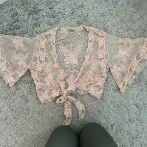 Rosa spetsbolero med fjärilsmönster - Säljer en söt rosa spetsbolero med vackert fjärilsmönster. Den har vida trekvartsärmar och knytning framtill. Perfekt för att lägga till en söt touch till din outfit.