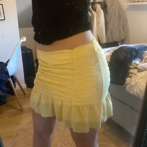 Gul kjol från Gina Tricot - Säljer en söt gul kjol från Gina Tricot med volangdetaljer och rynkad design. Kjolen är kort och perfekt för sommarens varma dagar. Tillverkad i ett lätt och bekvämt material.