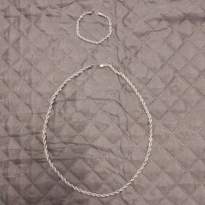 Silverfärgat halsband och armband - Säljer ett elegant set med ett silverfärgat halsband och matchande armband. Båda smyckena har en tvinnad design som ger en stilren och tidlös look. Perfekt för att komplettera din outfit med en touch av elegans.