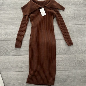 Brun ribbad klänning från Femme Luxe - Snygg brun ribbad klänning från Femme Luxe med en unik offshoulder-design. Klänningen har långa ärmar och en figurnära passform som framhäver silhuetten. Perfekt för en stilren look.