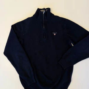 Mörkblå tröja med dragkedja från Gant - Säljer en stilren mörkblå tröja från Gant med halv dragkedja och broderad logga på bröstet. Tröjan har långa ärmar och ribbade muddar vid ärmslut och nederkant. Perfekt för en avslappnad men snygg look.