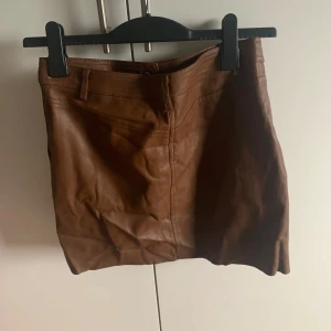 Brun skinnkjol med knappar - Snygg brun skinnkjol med knappar framtill. Perfekt för en trendig look. Kjolen har en kort längd och en stilren design.