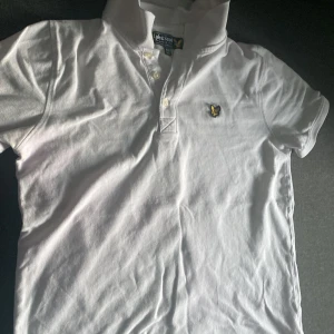 Vit kortärmad t-shirt från Lyle & Scott - Säljer en stilren vit kortärmad t-shirt från Lyle & Scott. Den har en klassisk krage och knappar framtill. Tillverkad i 100% bomull för en skön känsla. Perfekt för en avslappnad stil.