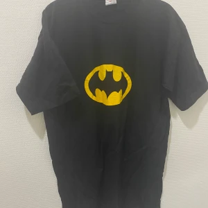Svart t-shirt med Batman-logga - Snygg svart t-shirt med den ikoniska Batman-loggan i gult på bröstet. Perfekt för alla superhjältefans! T-shirten har en klassisk passform och korta ärmar. Stlk kvar L och XL , 100% bomull 