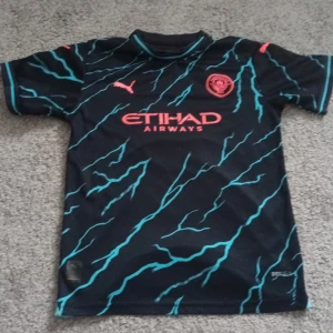 Svart fotbollströja med turkost mönster - Säljer en svart fotbollströja från Puma med turkost blixtmönster och röda detaljer. Tröjan har korta ärmar och texten 'DE BRUYNE 17' på ryggen. Perfekt för fotbollsfans! ⚽