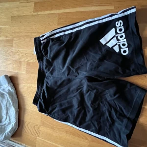 Adidas shorts - Säljer ett par svarta Adidas shorts med vita ränder längs sidorna och en stor logga på ena benet. Perfekta för träning eller avslappnade dagar. Elastisk midja för bekväm passform.