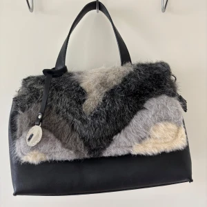 Svart handväska med pälsdetaljer - Unik svart handväska med mjuka pälsdetaljer i grå och beige toner. Medelstor väska med flera fickor. Handväskan kommer med extra axelband om man skulle vilja göra den längre.