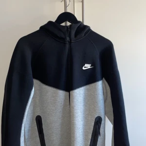 Svart och grå hoodie från Nike - Snygg hoodie från Nike i svart och grått med dragkedja och huva. Den har en klassisk design med Nike-loggan på bröstet och praktiska fickor framtill. Perfekt för en avslappnad stil.