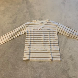 Randig långärmad tröja - Säljer en stilren randig tröja i beige och vit. Tröjan har långa ärmar och en klassisk rund halsringning. Perfekt för en avslappnad look.