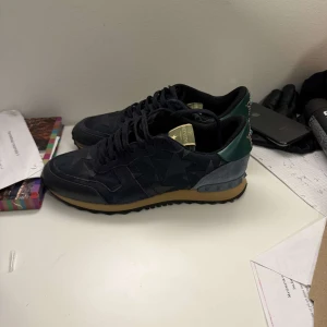 Valentino rock runners - Snygga Valentino Garavani sneakers i en mörk färgskala med stjärndetaljer på ovansidan. Skorna har en kontrasterande grön häl och en gummisula i brunt. Perfekta för en stilren look med en touch av lyx. Skick 9/10 och äkthets bevis finns. Priset är ej hugget i sten vid snabb affär!