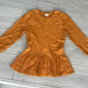 Orange stickad tröja från H&M - Säljer en snygg orange stickad tröja från H&M med en peplumdetalj i nederkant. Tröjan har långa ärmar och knappar längs ryggen för en unik look. Perfekt för att liva upp garderoben med lite färg!
