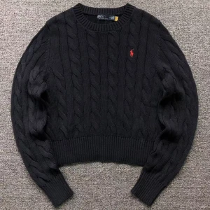 Svart stickad tröja från Ralph Lauren - Säljer en snygg svart stickad tröja från Ralph Lauren med kabelstickat mönster och röd logga på bröstet. Tröjan har långa ärmar och ribbstickade muddar vid ärmslut och nederkant. Perfekt för kyliga dagar!